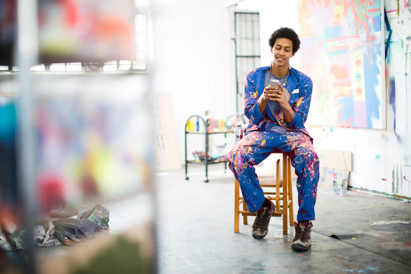 artiste, atelier, peinture, smartphone, homme, créatif, bleu, assis, sourire, multicolore, tabouret, studio, toile, vêtement, bombe, décontracté, intérieur, jeune, fenêtre, éclaboussure